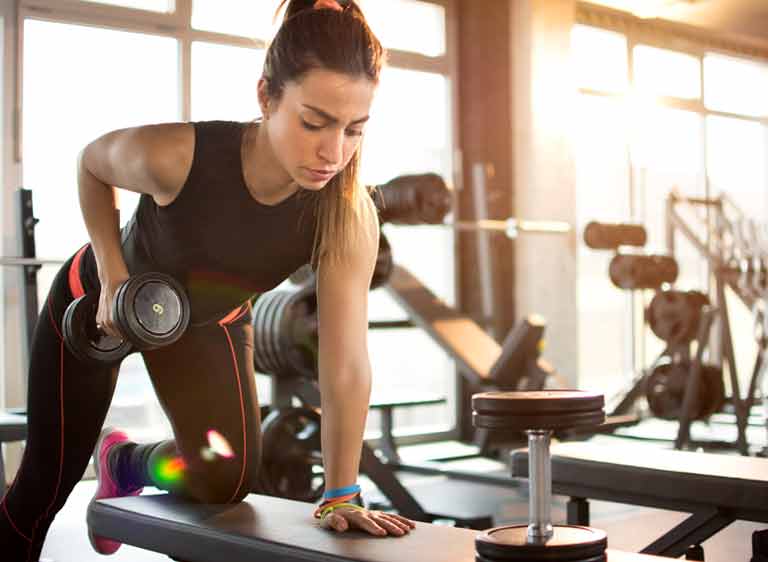 A woman does basic bench press exercises with a weight on one hand while holding with another hand on top off a bench at a gym.