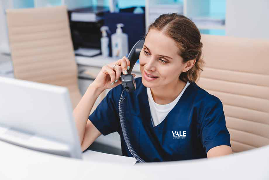 A female staff holds the phone to her ear while explaining insurance benefits to a patient.