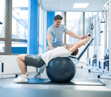 A male physical therapist uses a fitness ball to help a male patient with back stretching exercises.