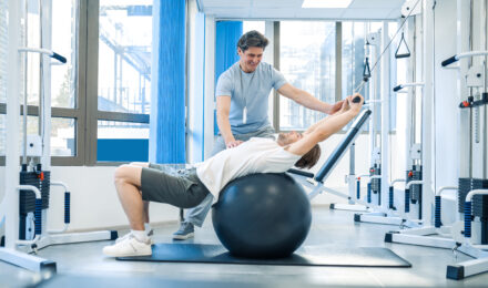 A male physical therapist uses a fitness ball to help a male patient with back stretching exercises.