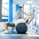 A male physical therapist uses a fitness ball to help a male patient with back stretching exercises.