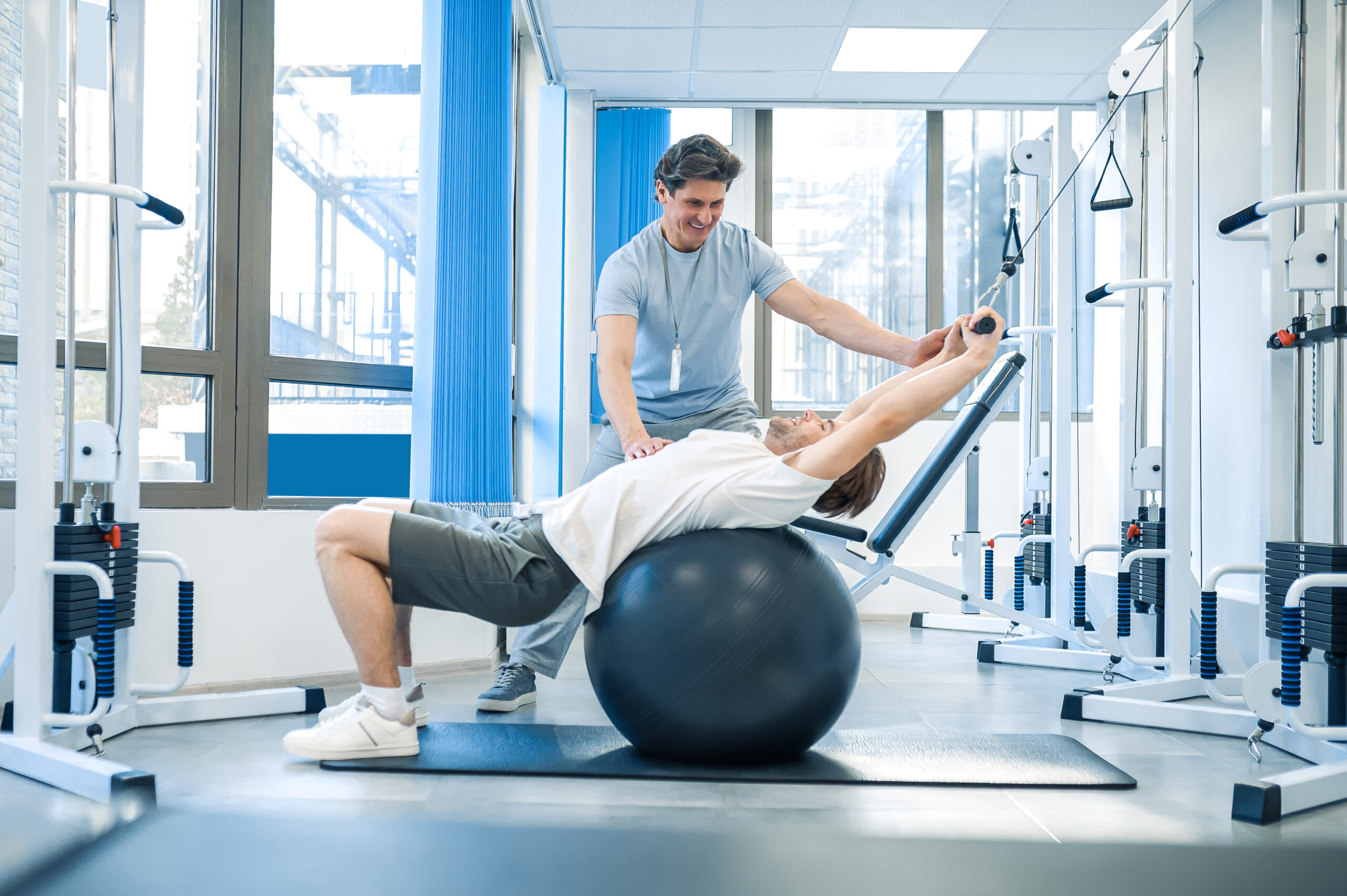 A male physical therapist uses a fitness ball to help a male patient with back stretching exercises.