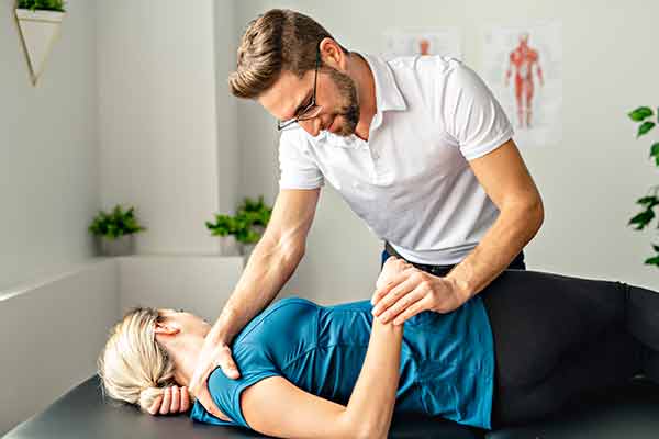 Male physical therapist moves female patient shoulder while holding her hand