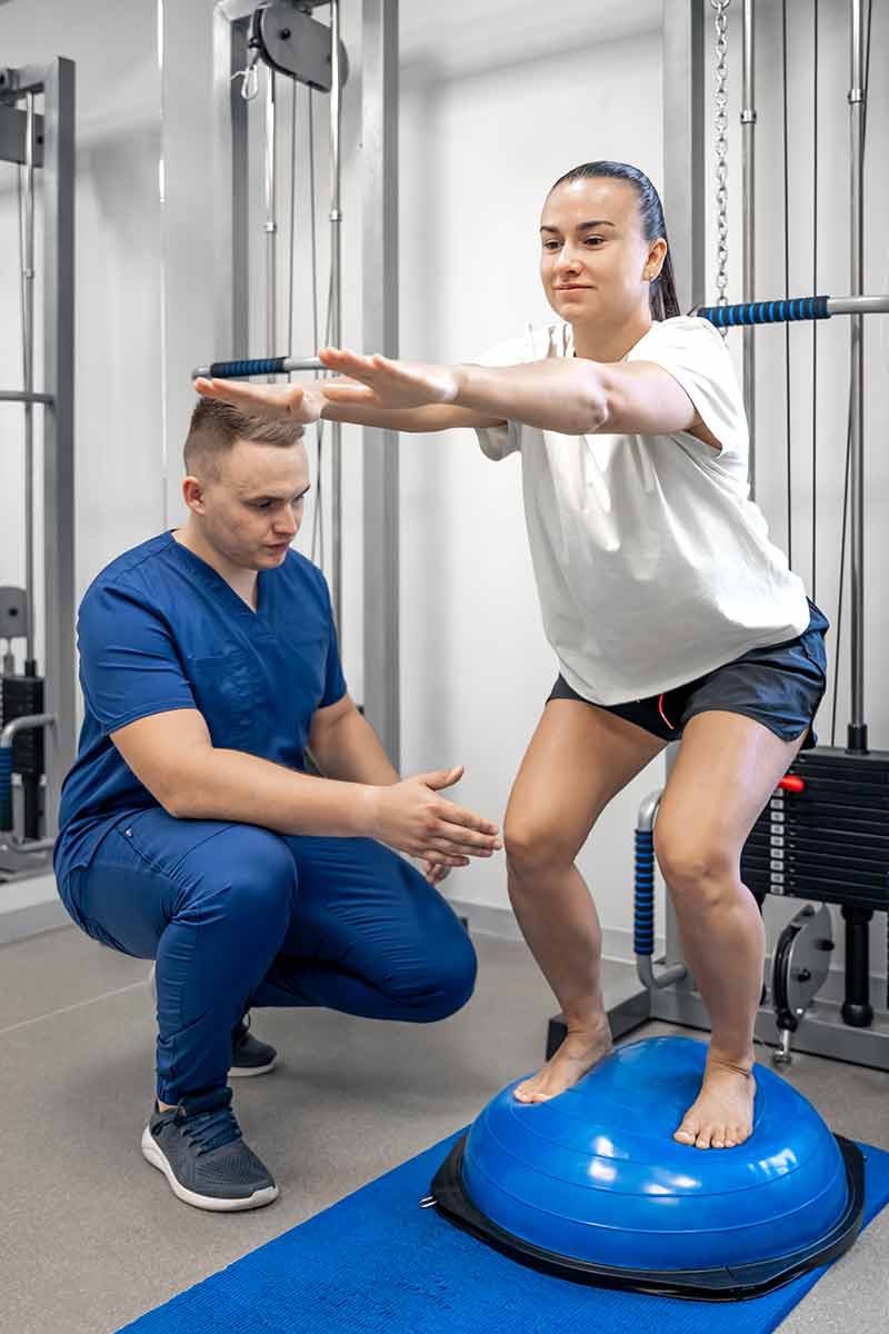 Woman doing post operative rehab balance exercises with a bosu ball under the assistance of physical therapist