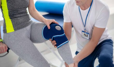 A male physical therapist holds the leg of a female patient with a knee compression sleeve.