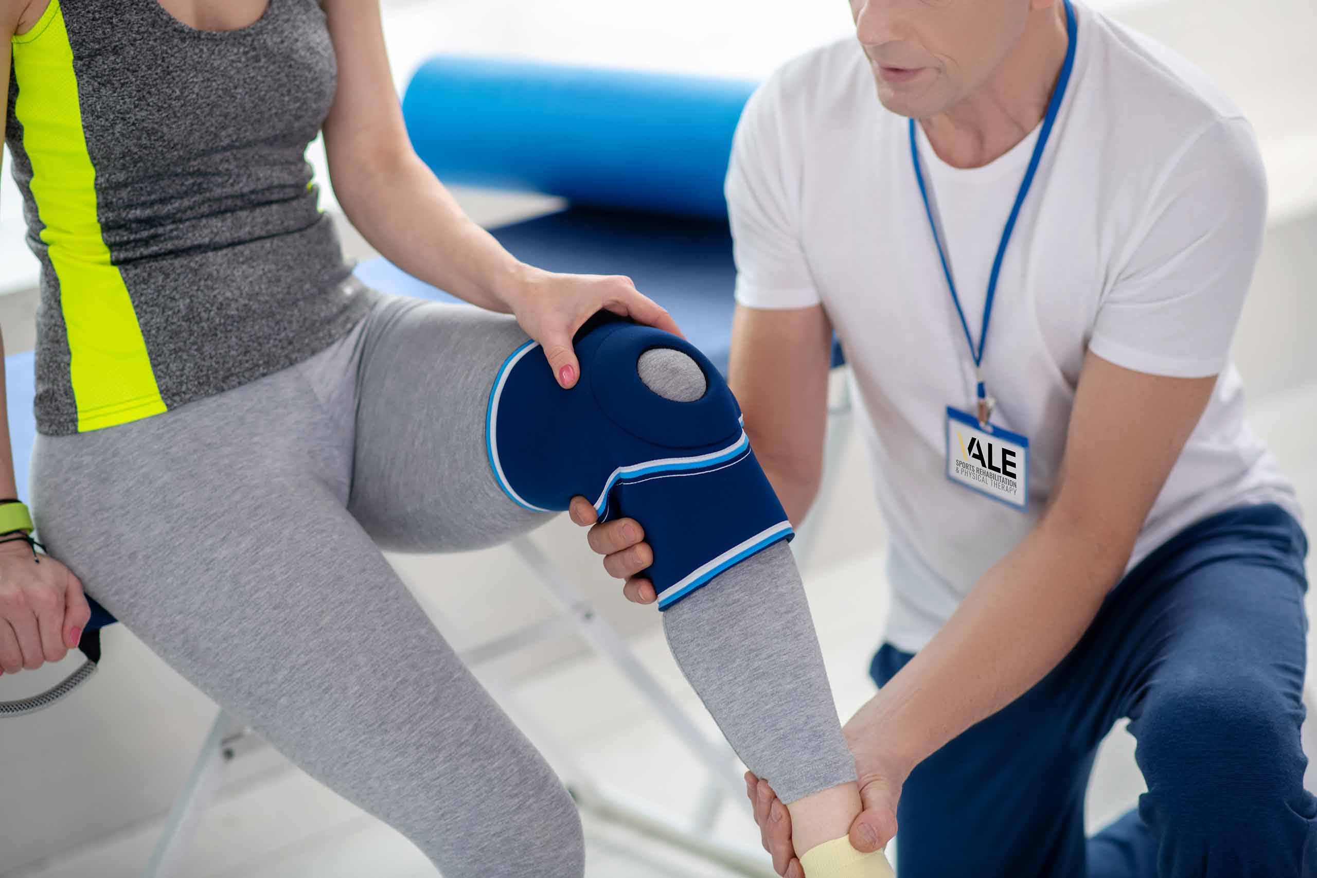 A male physical therapist holds the leg of a female patient with a knee compression sleeve.